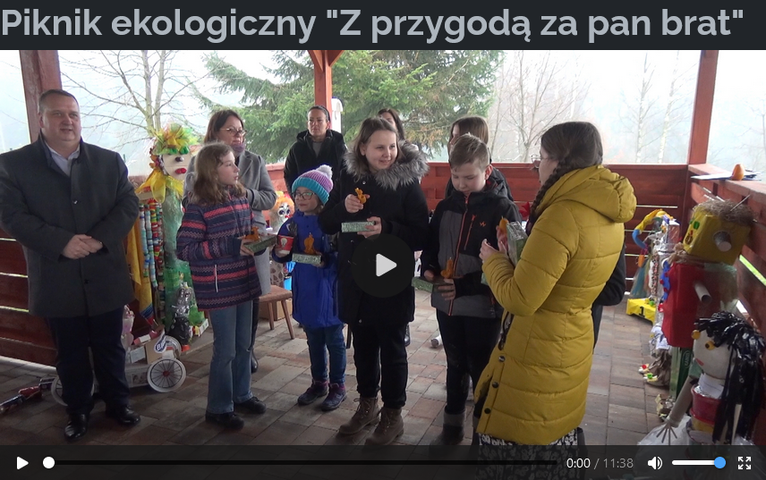 Zdjęcie jest zrzutem ekranu z wideo o tytule “Piknik ekologiczny "Z przygodą za pan brat”. Na zdjęciu widoczne są następujące elementy:  Kilka osób zgromadzonych pod zadaszeniem, różniących się wiekiem, w tym zarówno dzieci, jak i dorośli. Wokół ludzi widoczne są kolorowe dekoracje lub obiekty. Na środku znajduje się przycisk odtwarzania wskazujący, że jest to zatrzymany zrzut ekranu z wideo. Zdjęcie jest zarazem odnośnikiem do wideo na stronie http://itvpoludnie.pl/filmy/piknik-ekologiczny-z-przygoda-za-pan-brat.mp4 Atmosfera wydaje się swobodna i wspólna, sugerując spotkanie towarzyskie lub wydarzenie.