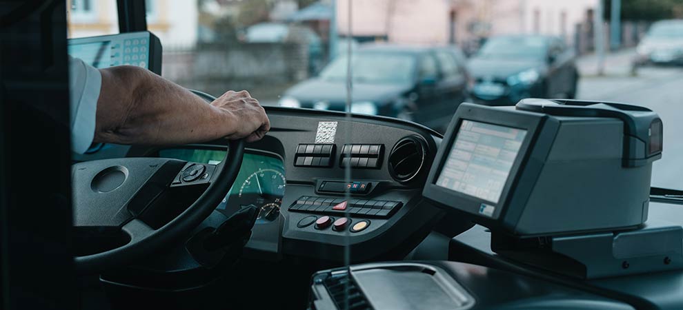 Obraz przedstawia wnętrze autobusu, z perspektywy kierowcy. Widać rękę osoby spoczywającą na kierownicy. Deska rozdzielcza zawiera różne przyciski. Na prawej stronie deski rozdzielczej zamontowany jest wyświetlacz cyfrowy, który wyświetla tekst i informacje na tle ciemnego tła. Przez przednią szybę widoczne są zaparkowane samochody i budynki, wskazując na miejskie otoczenie.